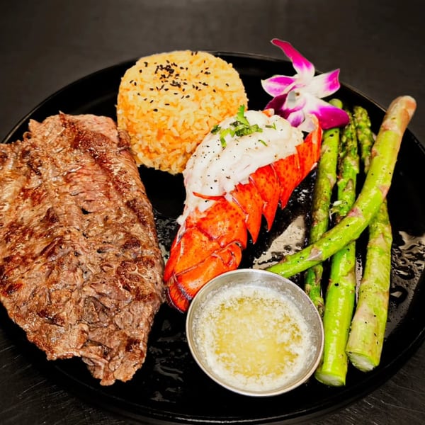 Grilled steak, lobster tail, rice, asparagus, and melted butter on a black plate.